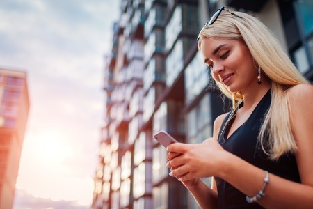 Young blonde businesswoman using smartphone by contemporary business center in city. Business, communication, technology and finance conceptの写真素材