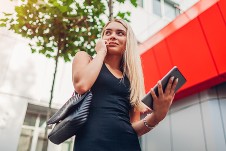 Young blonde businesswoman using smartphone and tablet by contemporary business center in city. Business, communication, technology and finance conceptの写真素材