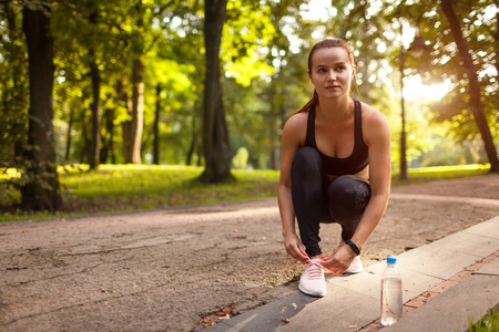 Young woman tying laces on her sneakers in summer park at sunset. Runner having rest. Sport concept.の写真素材