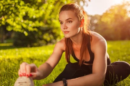 Young sportive woman doing exercises in summer park. Sportswoman stretching her legs sitting on grassの写真素材