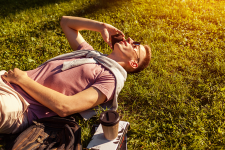Handsome college man chilling in spring campus park. Happy guy student lying on grass talking on smartphone with book and coffee besideの写真素材