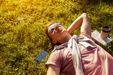 Handsome college man chilling in spring campus park. Happy guy student lying on grass with his phone, book and coffee beside at sunsetの写真素材