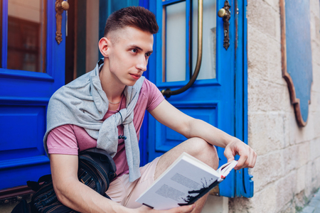 Handsome college man reading a book sitting by cafe. Happy guy student with backpack learning outdoors. High school educationの写真素材