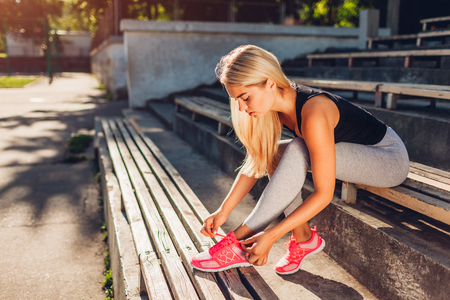 Young woman athlete tying sneakers sitting on bench on sportsground in summer. Preparation for trainingの写真素材