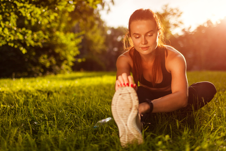 Young sportive woman doing exercises in summer park. Sportswoman stretching her legs sitting on grass. Sport conceptの写真素材