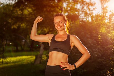 Young sportive woman showing biceps muscles in summer park. Strong happy girl smiling. Healthy lifestyle. Sport conceptの写真素材