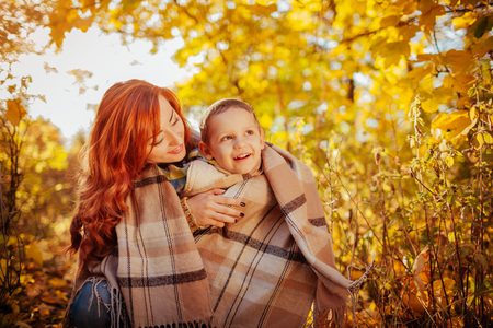 Happy mother and her little son walking and having fun in autumn forest. Woman wraps her kid in a scarfの写真素材