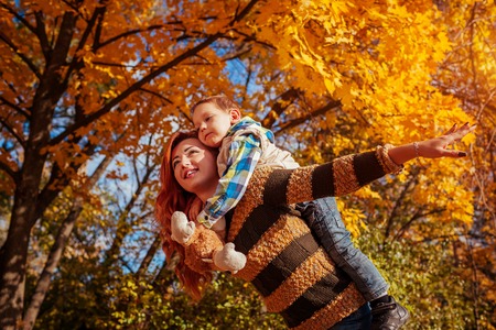 Happy mother and her little son walking and having fun in autumn forest. Kid riding on mothers backの写真素材