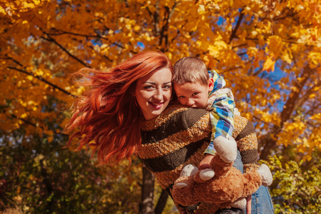 Happy mother and her little son walking and having fun in autumn forest. Kid riding on mothers backの写真素材