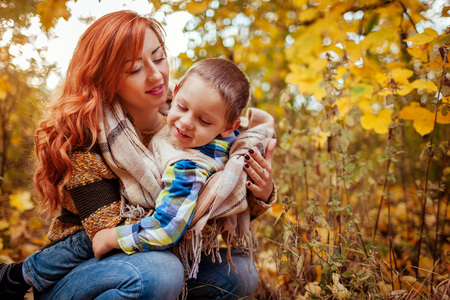 Happy mother and her little son walking and having fun in autumn forest. Woman wraps her kid in a scarfの写真素材