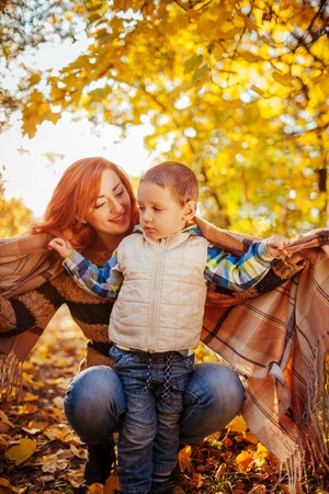 Happy mother and her little son walking and having fun in autumn forest. Woman wraps her kid in a scarfの写真素材