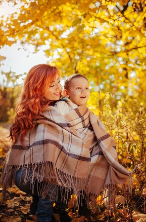 Happy mother and her little son walking and having fun in autumn forest. Woman wraps her kid in a scarfの写真素材