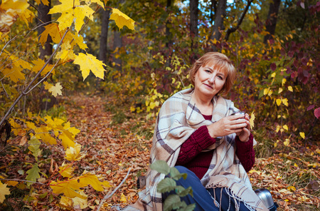 Happy middle-aged woman drinks tea in autumn forestの写真素材