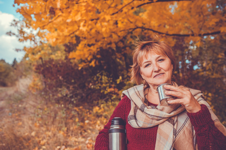 Happy middle-aged woman drinks tea in autumn forestの写真素材