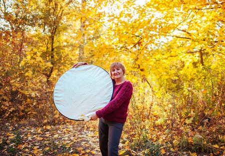 Middle-aged woman assists holding a reflector in autumn forest. Happy senior ladyの写真素材