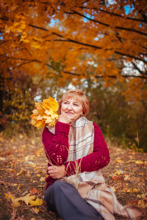 Middle-aged woman surrounded with yellow trees relaxes in autumn forest. Lady enjoying nature holding bouquet of leavesの写真素材