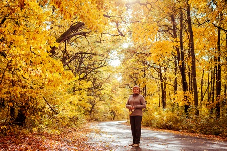 Middle-aged woman surrouded with yellow trees walks in autumn forest. Happy senior lady enjoying natureの写真素材
