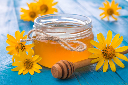 Honey in a glass jar with stick surrounded with yellow flowers on blue wooden tableの写真素材