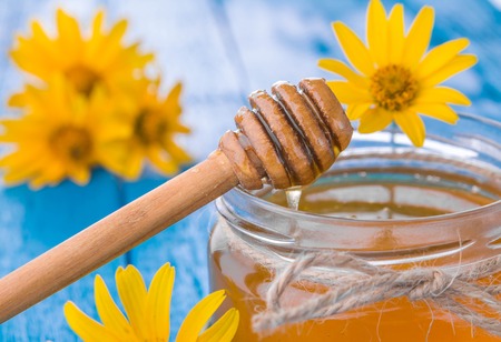 Honey in a glass jar with stick surrounded with yellow flowers on blue wooden tableの写真素材
