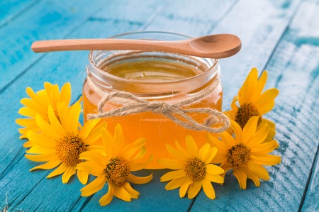 Honey in a glass jar with stick surrounded with yellow flowers on blue wooden tableの写真素材