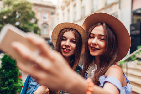 Outdoor portrait of two young beautiful fashionable women taking selfie using phone. Happy girls having fun in city. Best friends wearing hatsの写真素材