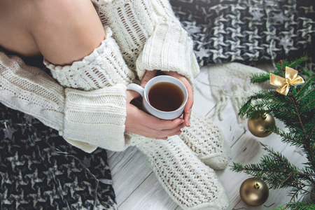 Woman holds a cup of tea beside her legs under Christmas tree