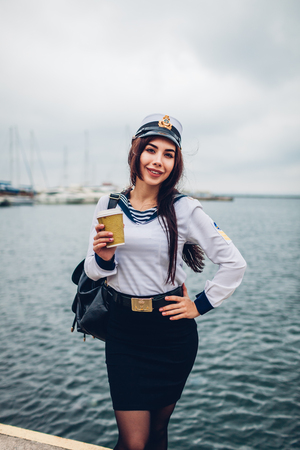 College woman student of Marine academy drinking coffee by sea wearing uniform. Girl walking in seaport of Odessa on pierの写真素材