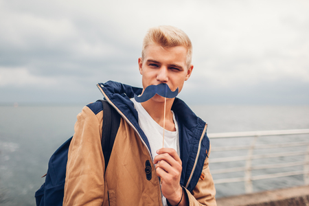 happy college student with backpack holding photo booth props moustache by sea. Guy having fun outdoors. Man winkingの写真素材