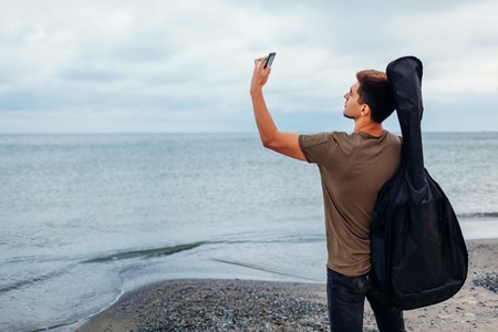 Young man with acoustic guitar in case taking selfie using phone on cloudy beach