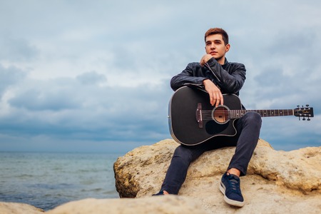 Young man with acoustic guitar sitting alone on beach surrounded with rocks on rainy dayの写真素材