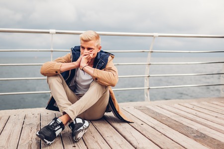 Upset young man sitting by sea on pier in Odessa thinking on rainy autumn day. Depression and stress concept. Mental healthの写真素材