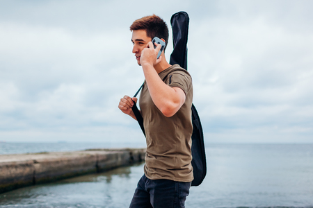 Young man carrying acoustic guitar in casw and talking on phone walking on cloudy beachの写真素材