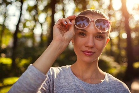 Smiling young woman taking off her sunglasses in park. Beautiful girl with natural makeup looking at camera outdoorsの写真素材