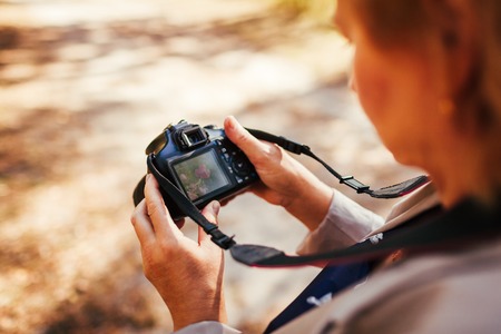 Middle-aged woman checking images on camera in autumn forest. Stylish senior woman walking and enjoying hobby shooting photosの写真素材