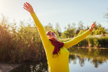 Middle-aged woman feeling free and happy on river bank on autumn day. Senior lady raising hands up. Harmony and balance with natureの写真素材