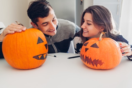 Young couple making jack-o-lantern for halloween on kitchen. Happy man and woman comparing their pumpkinsの写真素材