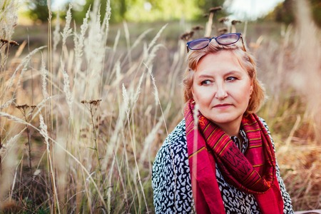 Stylish middle-aged woman chilling in autumn field. Senior lady wearing clothes and accessories....