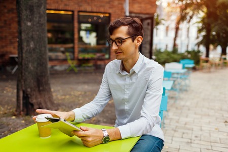 Young businessman enjoys coffee in outdoor cafe using tablet. Handsome guy drinking tea on city street holding tabletの写真素材
