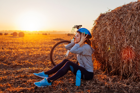 Young bicyclist having rest after a ride in autumn field at sunset. Woman taking off helmet sitting by haystack. Sport recreation conceptの写真素材