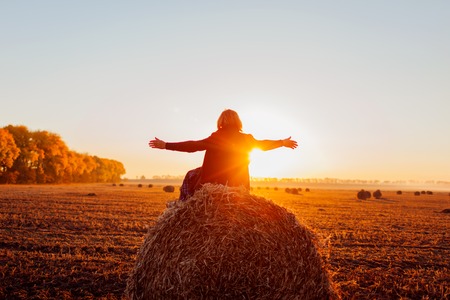 Happy middle-aged woman sitting on haystack in autumn field and feeling free with arms opened. Relaxing and admiring nature at sunsetの写真素材