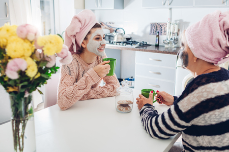 Mother and her adult daughter drinking tea with facial masks applied. Women chilling and talking on kitchen. Family valuesの写真素材
