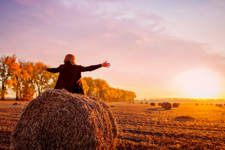 Happy middle-aged woman sitting on haystack in autumn field and feeling free with arms opened. Relaxing and admiring nature at sunsetの写真素材