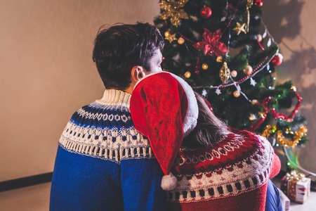 Couple in love sitting next to a Christmas tree, wearing Santa's hat and hugging. Young people celebrating New year at home at nightの写真素材