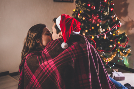 Couple in love sitting next to a Christmas tree, wearing Santa's hat and hugging. Young people celebrating New year at home at nightの写真素材