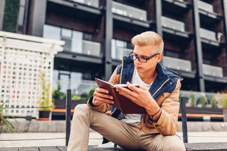 College student with backpack reading book chilling by modern hotel on rainy autumn day. Education conceptの写真素材