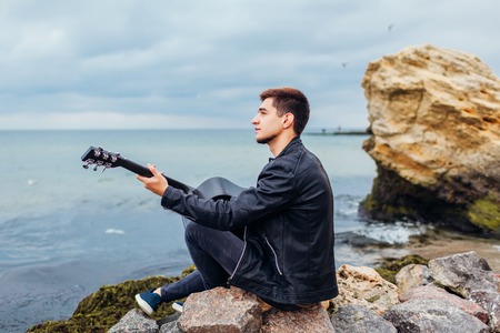 Young man with acoustic guitar playing alone on beach surrounded with rocks on rainy dayの写真素材