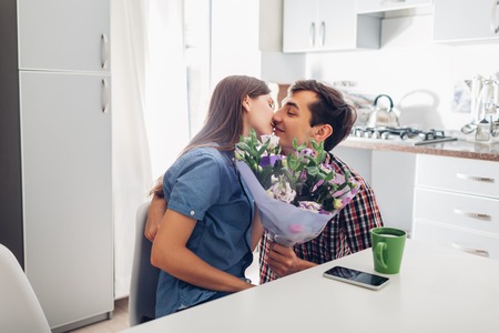 Young man gifting bouquet of flowers to his girlfriend in kitchen. Happy couple hugging. Romantic surprise at homeの写真素材