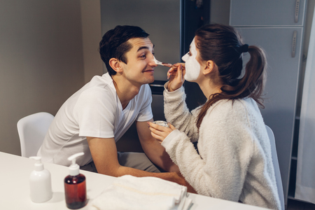 Woman applying clay mask on her boyfriends face. Young loving couple taking care of skin at homeの写真素材