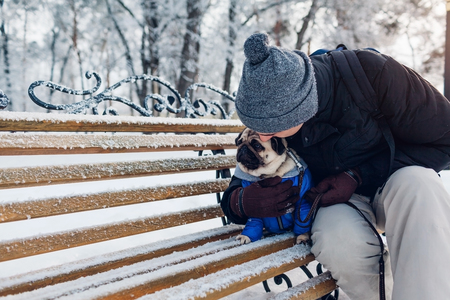 Pug dog sitting on bench with his master. Puppy wearing winter coat. Man hugging his pet in winter park. Best friendの写真素材