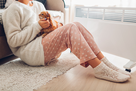 Using heater at home in winter. Woman warming body sitting by device with cat and wearing warm clothes. Heating season.の写真素材
